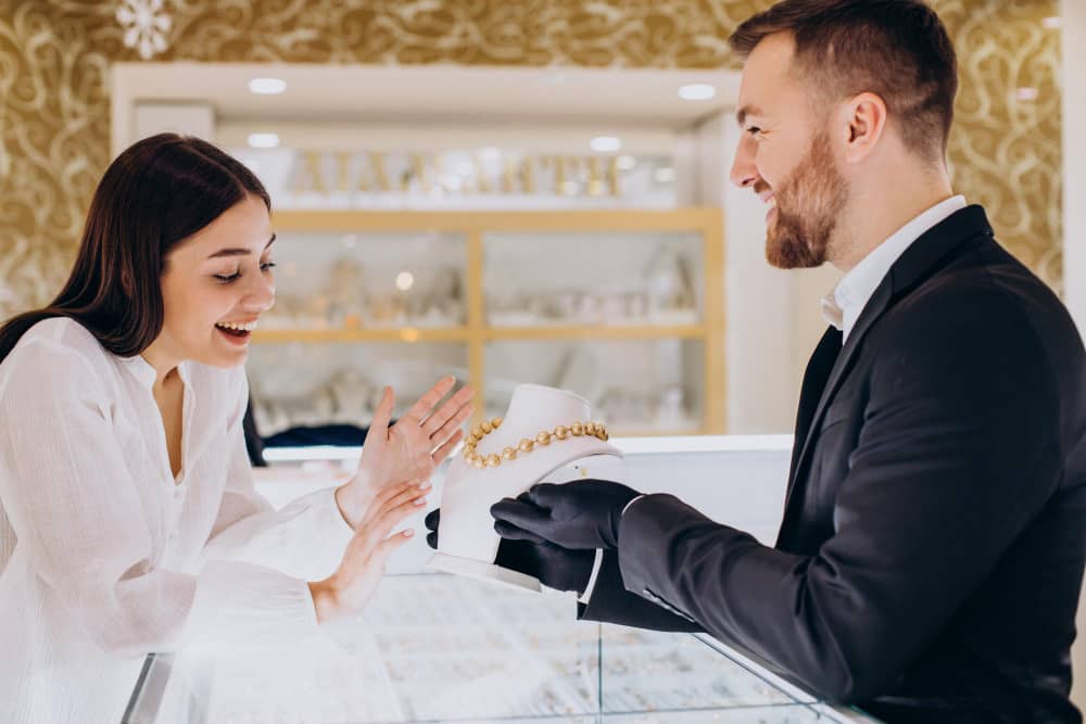 Un bijoutier en costume présente un collier en perles dorées à une jeune femme ravie.